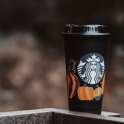 black Starbucks cup with pumpkins on it sits on top of a granite counter; there is a very blurry brownish background.