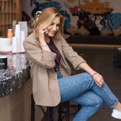 woman wears blazer with jeans; she is sitting on a stool at a coffee shop, talking on her cell phone, and laughing