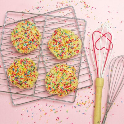 sugar cookies with lots of sprinkles sit on baking racks with a heart-shaped whisk next to the rack; there is a pink background.