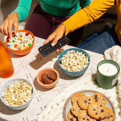 A coffee table with snacks, a bottle of wine, and a candle; two people's hands are visible, one holding a TV remote