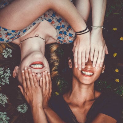 two women smiling and laying down in opposite directions; each woman covers the other woman's face with her hands.
