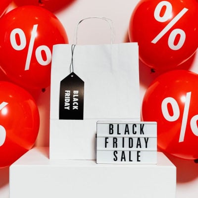 adjustable board reads "BLACK FRIDAY SALE;" it sits in front of a white gift bag with a black tag hanging off it that reads "BLACK FRIDAY." In the background are red balloons with percentage signs.