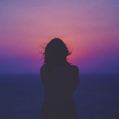 woman stands silhouetted against a sunset with oranges, pinks and purples; she has her arms wrapped around herself and is facing away from the camera