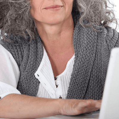 woman with gray hair works at a computer; she wears a crisp white blouse with a gray sweater over her shoulders