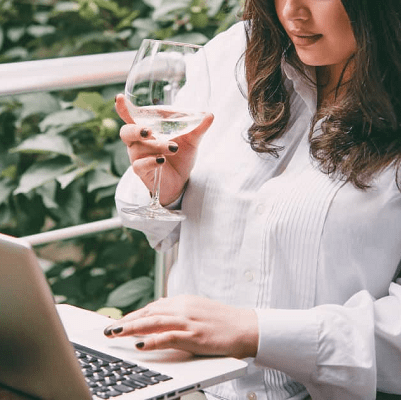 stylish professional woman wears plus-size white blouse, sips wine, and shops on her computer while sitting on a balcony