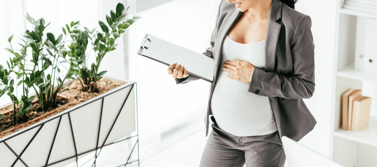 pregnant professional woman wearing a gray suit holds a clipboard; one hand is on her pregnant belly